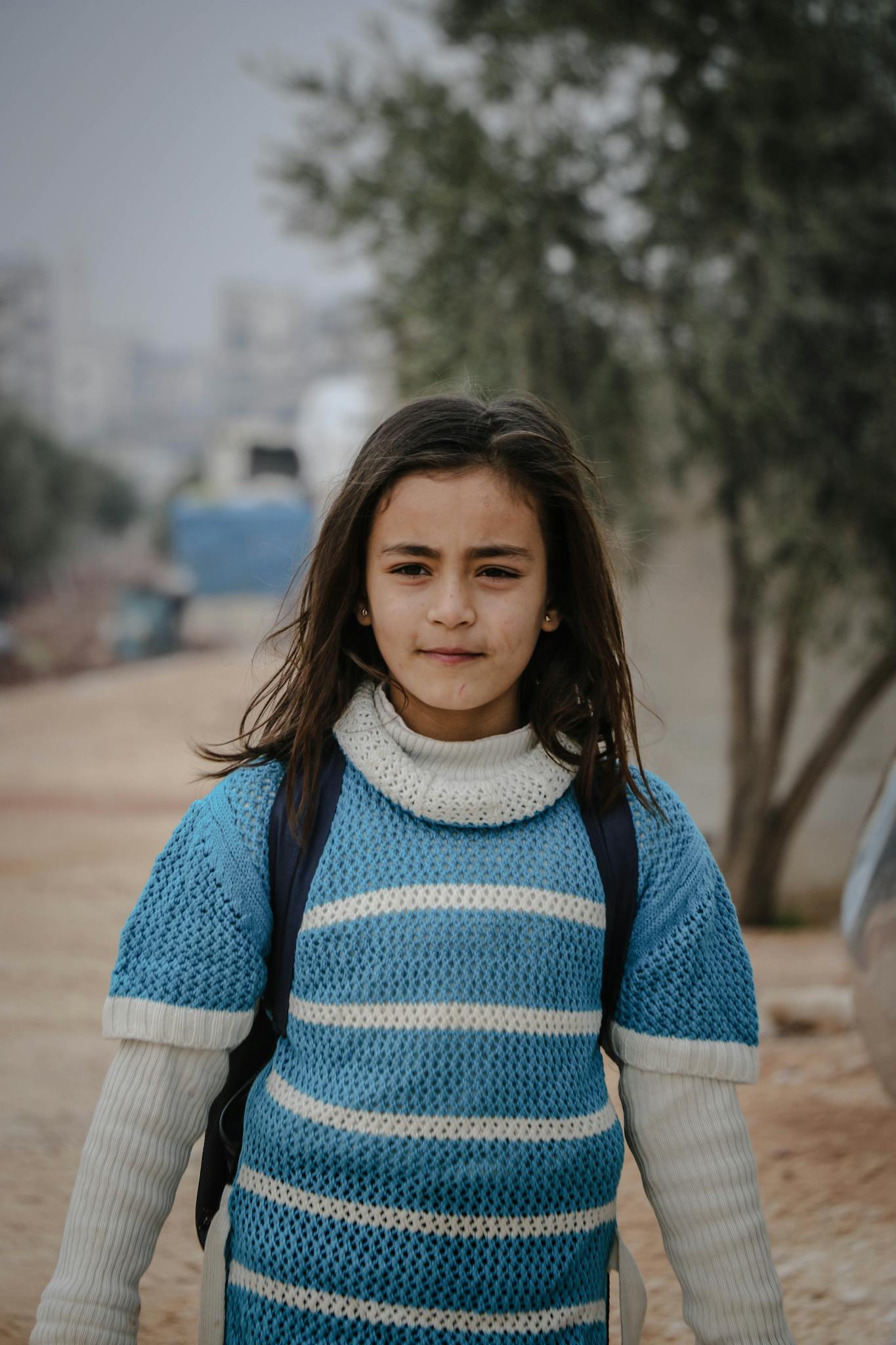 A young girl wearing a knitted sweater stands outdoors in Idlib, Syria, with a neutral expression.