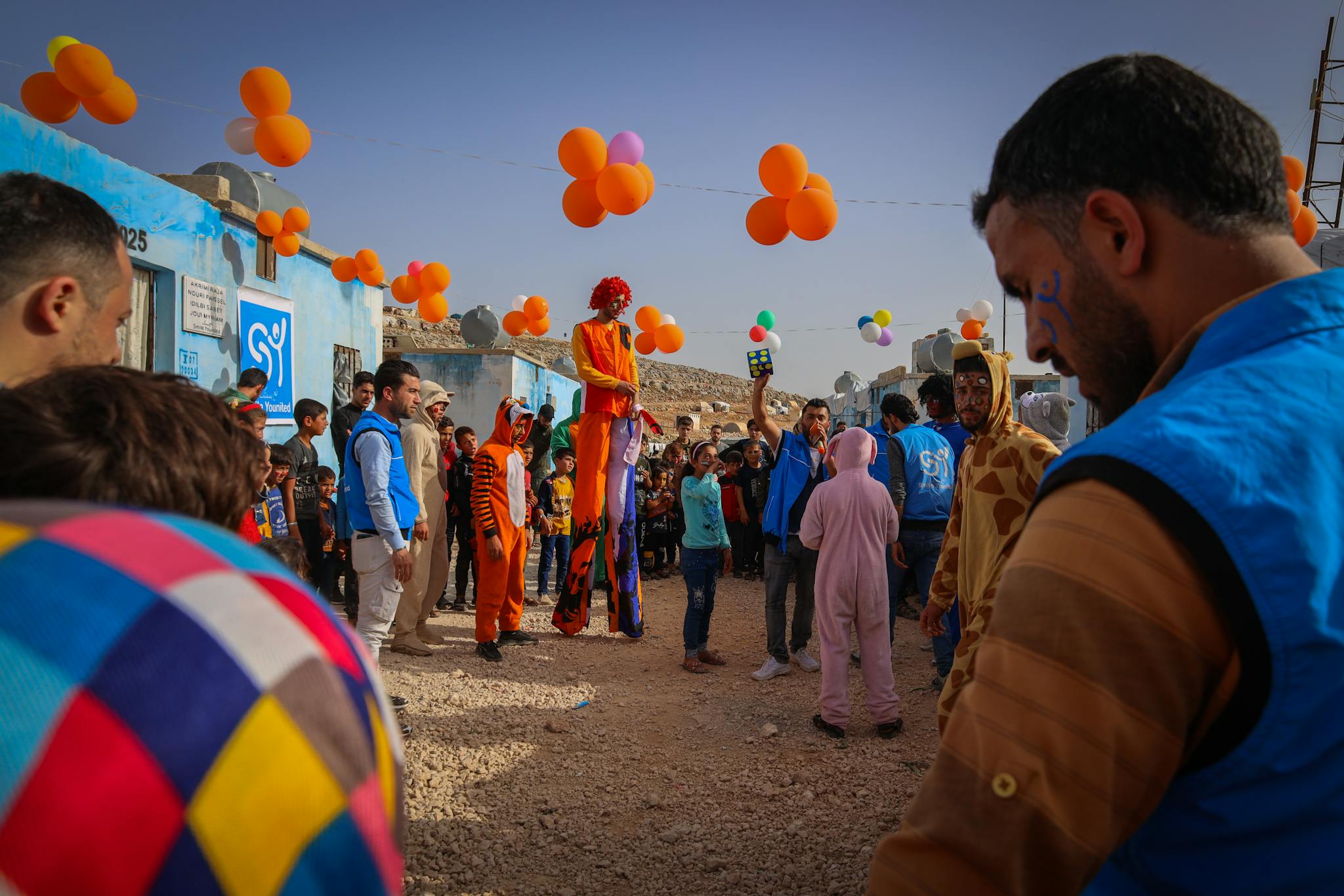 Colorful children's party outdoors featuring clowns, costumes, and balloons in Idlib, Syria.